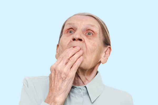 Lifestyle portrait of an elderly woman with a hand covering her mouth on a blue background. Surprised old lady pensioner with a frightened expression on her face. Concept of emotional shock.