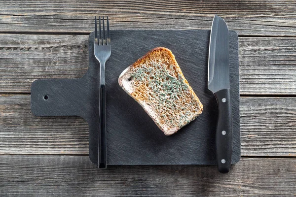 A piece of rye bread spoiled in mold is rotting on a black slate cutting board next to a serving knife and fork on a wooden table background. The concept of freeganism.