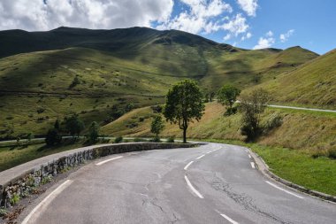 Yeşil tepelerle çevrili kıvrımlı bir yol ve beyaz bulutlu mavi bir gökyüzü. Col de Peyresourde, Fransa