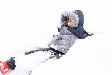 Mutlu çocuk tamamen karlı zeminde sırt üstü yatıyor. Çocuğun kafasında bir başlık ve kışlık bir ceket var..