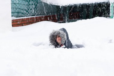 Mutlu çocuk tamamen karlı zeminde sırt üstü yatıyor. Çocuğun kafasında bir başlık ve kışlık bir ceket var..