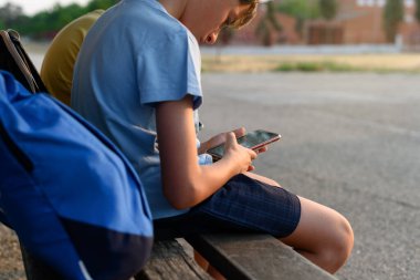 Sırt çantaları ve cep telefonlarıyla bankta oturan bir çocuk. Mobil cihaz bağımlılığı kavramı