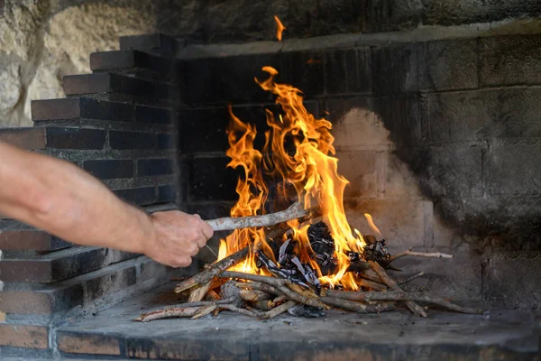 Man's hand throws a branch into a burning fireplace in a house. Rustic oven with burning trunks and branches. Energy saving concept