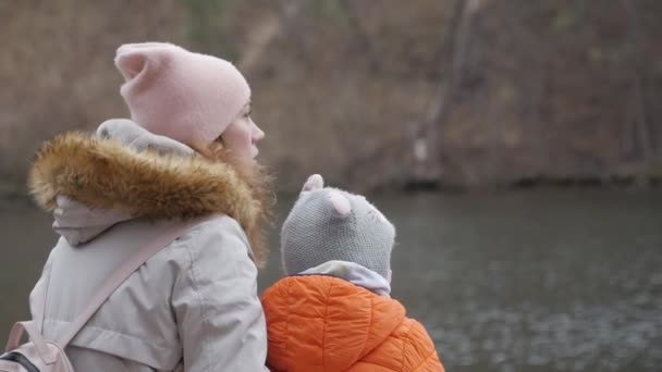 Une mère et une petite fille s'assoient sur la rive de la rivière sur le banc et parlent plus tard à l'automne.. — Video