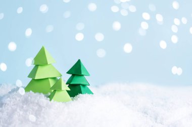 Noel ve kış kartı, karla kaplı bir tepenin üzerinde üç yeşil kağıt ağacı, bokeh ışıkları, kağıt işçiliği.