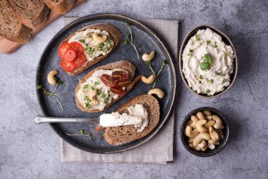 Mayalanmış fındıklı kase, kaju peynirli dilimlenmiş ekmek, domatesler ve gri arka planda mikro yeşillikler..
