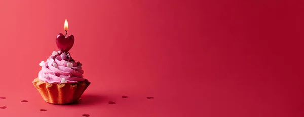 Valentine's day, cupcake with cream and burning heart shaped candle on red background, banner.