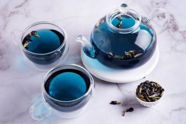 Kelebek çiçekli iki bardak ve cam çaydanlık çay ve mermer arka planda kurutulmuş klitorya çiçeği, mavi anchan bitkisel içeceği..