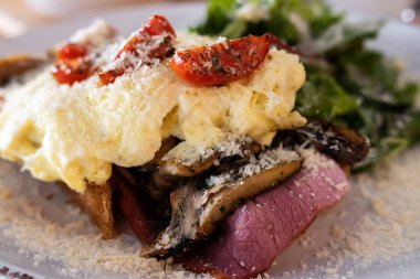 Close-up of breakfast sandwich with toasted bread, ham, mushrooms, scrambled egg and tomato.