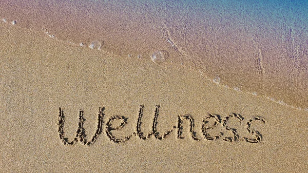 Word Wellness handwritten on the sand. Beach and soft wave background.