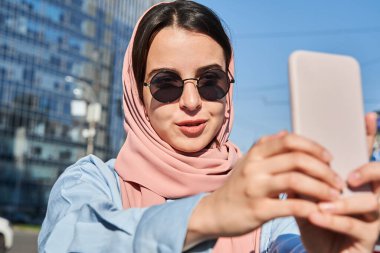 Başörtüsü takan kadın seyahat ederken cep telefonuyla selfie çekiyor.