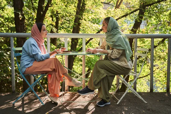 Mujer musulmana en hiyab sentada con comida en la cafetería de la calle y mirando a su mejor amiga —  Fotos de Stock