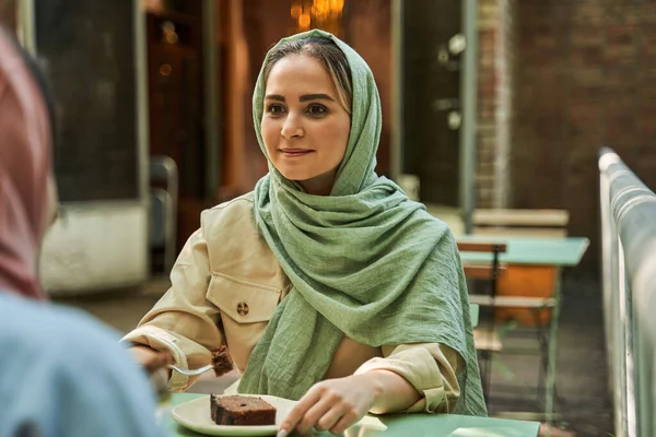 Mujer tranquila en hijab mirando a su mejor amiga con sonrisa amistosa mientras comen juntos —  Fotos de Stock