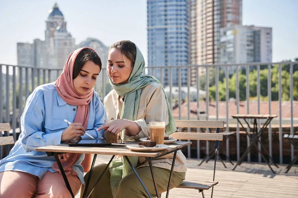 Két lány abayát visel, akik figyelmesen dolgoznak a tabletnél, miközben a kávézóban töltik az idejüket. — Stock Fotó