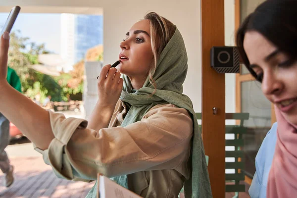 Mujer musulmana mirando la cámara frontal de su smartphone y aplicándose tinte en los labios —  Fotos de Stock