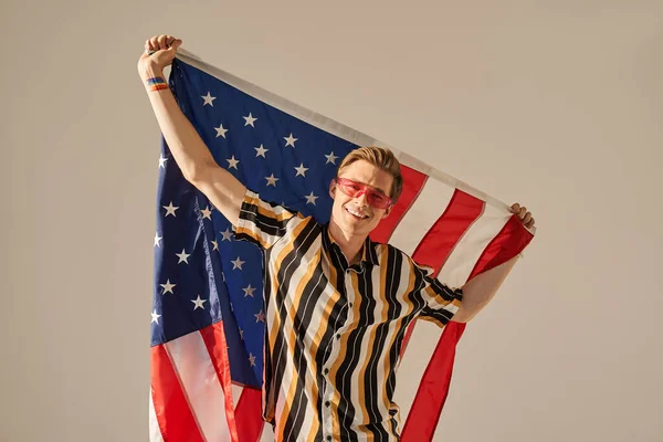 Hombre con el símbolo del arco iris en su brazo sosteniendo la bandera de los estados unidos y sonriendo feliz — Foto de Stock