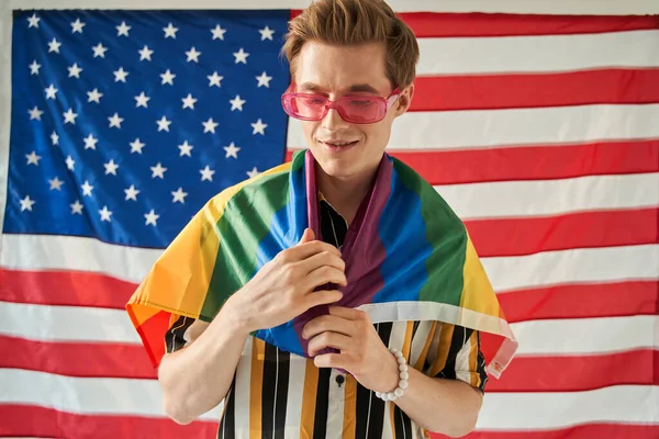 Feliz hombre bisexual orgulloso está envolviendo en arco iris bandera de color LGBT en el fondo de EE.UU.. — Foto de Stock