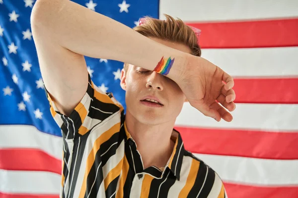 Moderno chico americano con símbolo lgbt en su muñeca ocultando sus ojos detrás de su mano — Foto de Stock