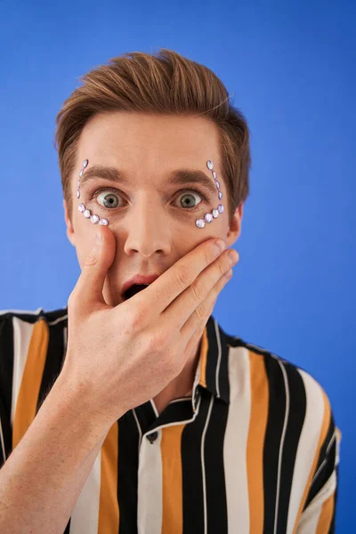 Mann mit Strass im Gesicht steht und versteckt mit der Hand seinen offenen Überraschungsmund — Stockfoto