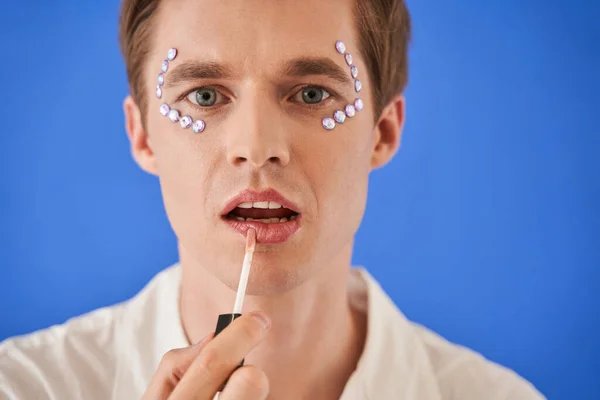 Hombre con pedrería bajo los ojos aplicando brillo labial y mirando atentamente a la cámara — Foto de Stock