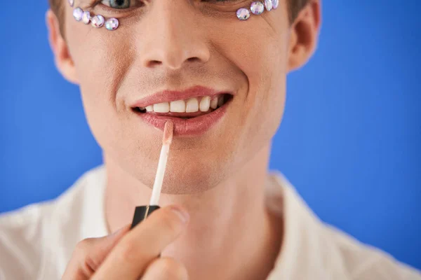 Bisexual hombre aplicando maquillaje en sus labios aislado sobre el vibrante fondo de color azul — Foto de Stock