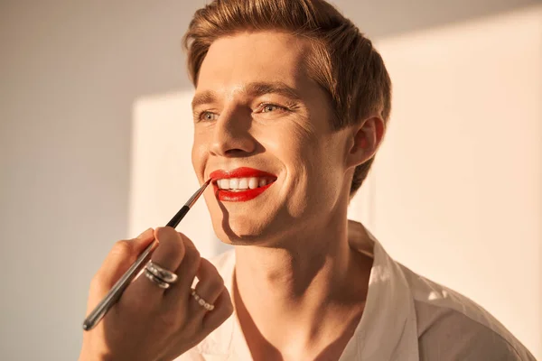 Hombre sonriendo mientras visagiste femenino aplicando lápiz labial rojo brillante en su boca — Foto de Stock