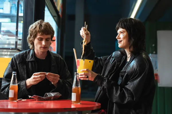 Flicka äter nudlar medan hennes pojkvän rullar en cigarett på kvällsgatan café — Stockfoto