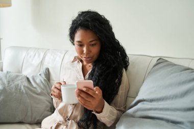 Kadın kahve içiyor ve evde vakit geçirirken akıllı telefonuna bakıyor.