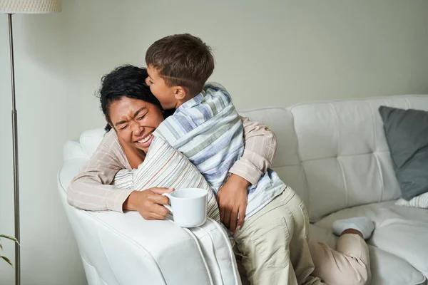 Милый маленький мальчик обнимает свою прекрасную мать, сидя на диване — стоковое фото
