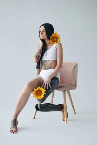 Татуированная девушка с протезной ногой держит подсолнухи и смотрит в сторону, сидя — стоковое фото
