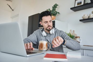 Bir beyefendi dizüstü bilgisayarda çalışırken saatlerine bakıyor ve sabah içkisinin tadını çıkarıyor.