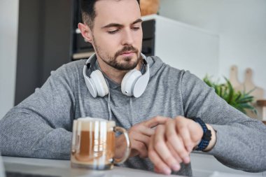 Mutlu beyaz erkek evde saatlere bakarken dizüstü bilgisayar kullanıyor.