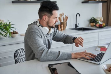 Evde dizüstü bilgisayar kullanırken kol saatine bakan traşsız, gülümseyen genç bir adam.