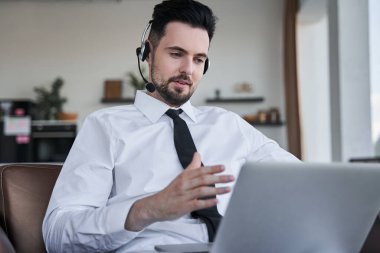 Kulaklık takan bir iş adamı koltuğunda oturmuş video görüşmesi yaparken el kol hareketi yapıyor.