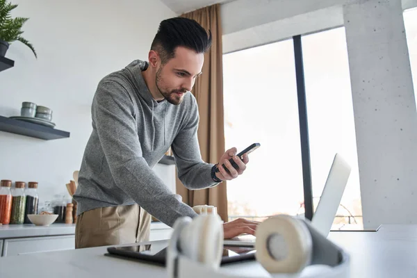 Hombre sosteniendo el teléfono inteligente mientras usa el ordenador portátil durante el trabajo de forma remota desde casa — Foto de Stock