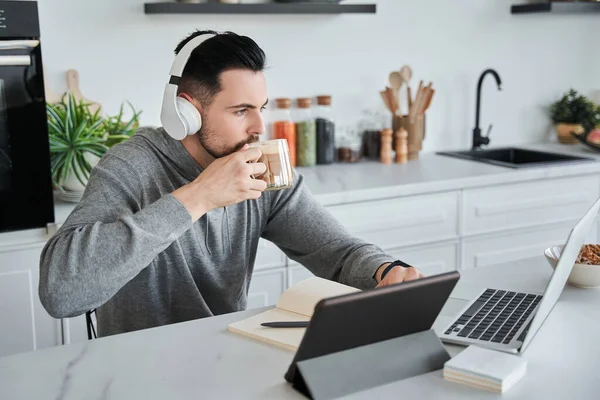 Kulaklık takan bir adam laptopun önünde oturup çalışırken kahve içiyor.