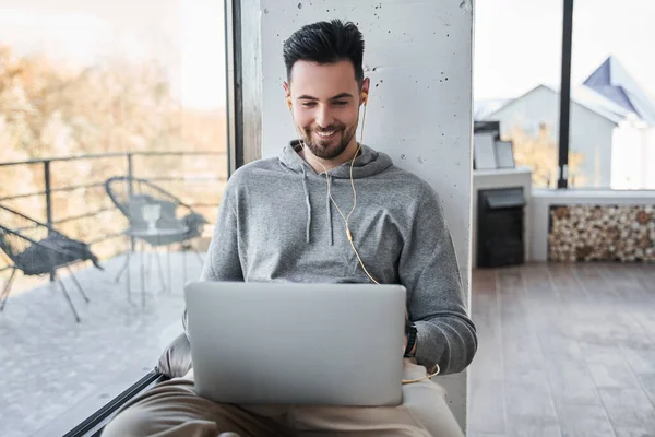 Homem sentado no peitoril da janela em casa e usando seu laptop com sorriso de prazer — Fotografia de Stock