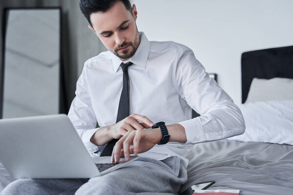 Man wearing shirt and tie looking at his wristwatch while sitting at the bed