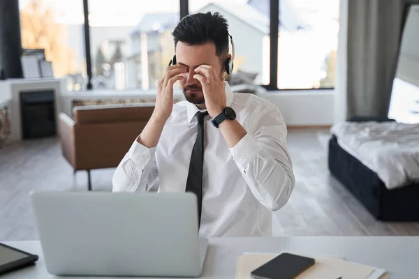 Kulaklık takan sakallı adam dizüstü bilgisayarın önünde otururken gözlerini ovalıyor. — Stok fotoğraf