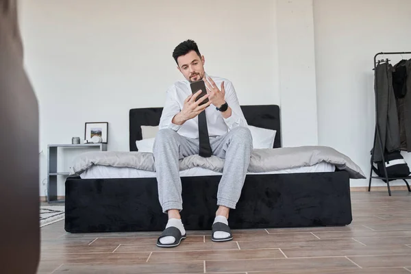 ビジネスマンは、ベッドに座って、彼のスマートフォンでオンライン通話を介してチャット — ストック写真