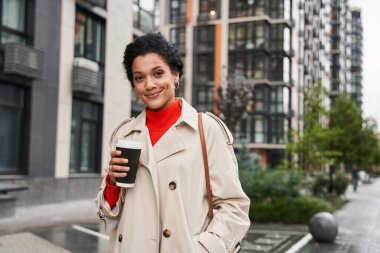 Elinde bir fincan kahve tutan ve kameraya mutlu bir ifadeyle bakan kadın.