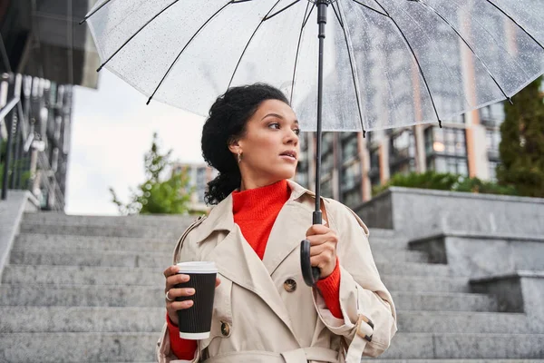 Ruhige brünette Frau läuft unter transparentem Regenschirm und versteckt sich vor dem Regen — Stockfoto