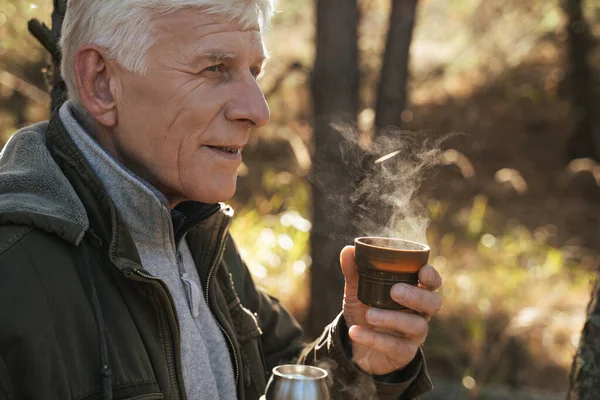 Positive tourist holding cup with hot tea while sitting on the ground in pinewood — Stock Photo, Image