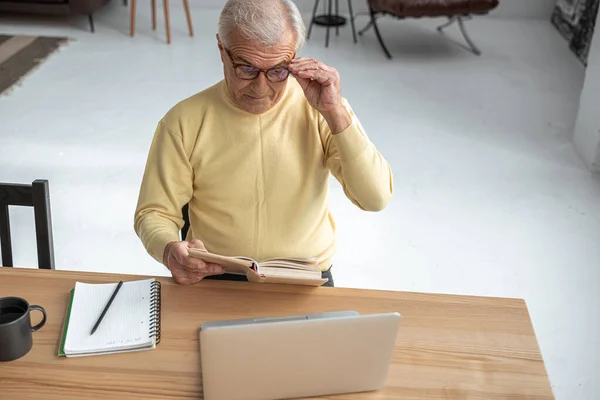 Homem maduro endireita óculos enquanto lê livro interessante com expressão séria — Fotografia de Stock