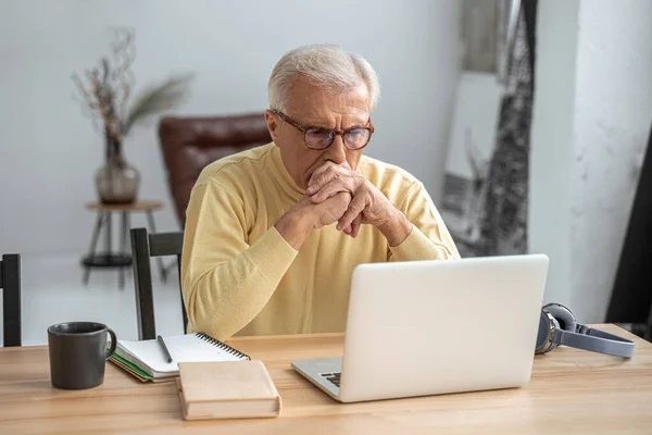 Старий в окулярах дивиться на екран ноутбука з серйозним виразом — стокове фото