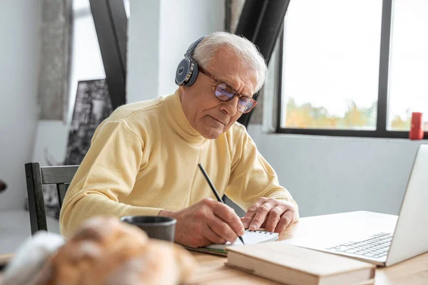 Homem maduro usando fones de ouvido usando laptop e escrevendo notas ao fazer chamada de vídeo — Fotografia de Stock