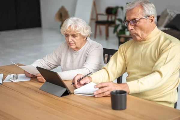 Pensiv koncentrerad äldre familj par sitter vid tablett och studera pappersräkningar — Stockfoto