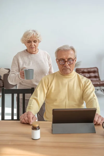 Kvinna som håller kopp med te medan hennes man har online samtal med läkare — Stockfoto