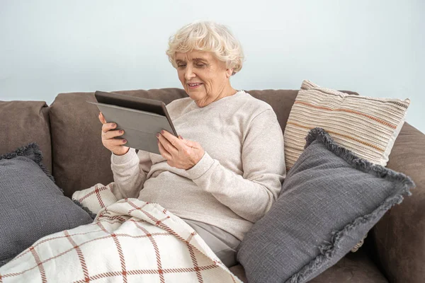 Mujer de edad utilizando la tableta digital mientras se relaja en el sofá y leer el libro electrónico —  Fotos de Stock