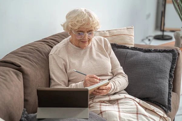 Mujer mayor escribiendo algo en su portátil mientras sostiene el dispositivo electrónico en el sofá — Foto de Stock